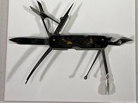 Black multi-tool with multiple tools unfolded, showing knife blade, scissors, file, pick, and other tools.