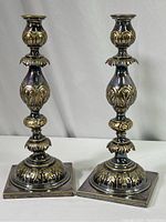 Pair of silverplate Shabbat candlesticks with floral patterns and square bases, on neutral surface.
