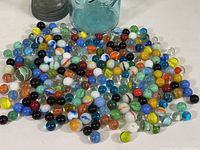 Large assortment of vintage glass marbles outside the jar, showing variety of colors and patterns, jar visible in background.