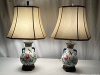 Pair of Chinese inspired porcelain table lamps with beige fabric lampshades lit against a white background.