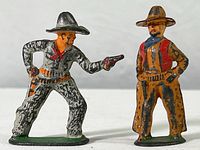 Two vintage hand painted metal cowboy toy figures, one aiming a pistol and one standing with hands on hips, showing front view.
