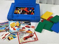 Blue plastic Lego carrying case open, filled with assorted vintage Lego pieces and instruction booklets spread in front
