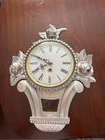 Antique wooden clock with a round face, Roman numerals, decorative leaf carvings and a reflective inset piece in the base.