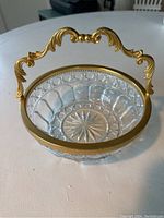 Photo of clear pressed crystal serving bowl with scalloped edge rim and starburst pattern base. Gilt metal trim around the rim and ornate gilt metal handle attached to the rim, featuring scroll details.