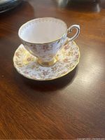 Photo of a delicate English bone china teacup and saucer set featuring 24K gold leaf decorations and gold trim on a wood surface.