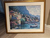 Frontal photograph of the framed giclee print showing the Mediterranean coastal scene with boats and houses, with lighting highlighting the colors and texture.