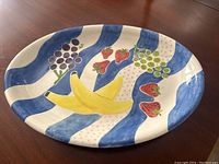 Oval ceramic platter showing hand painted fruit designs with bananas, strawberries, grapes, and green berries on a blue and white striped background.