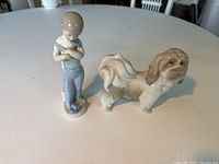 Two Lladro porcelain figurines placed upright on a round white table, showing front views of a small boy holding toys and a Lhasa Apso dog.