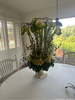 Photo of silk floral centerpiece on round white table near window with white chairs.