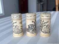 Front view of three cylindrical Asian-style plastic pill boxes arranged side by side showing detailed black line art of rural scenes and figures.