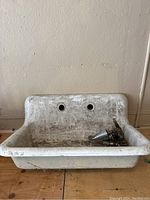 Front view of white cast iron farmhouse sink showing dual faucet holes, central drain, and attached faucet components. Sink surface shows heavy wear and dirt.