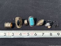 All 7 finger rings placed on a black background next to a ruler showing size between 2-9 inches, for overview and scale
