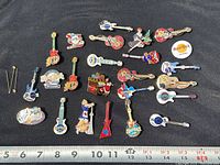 Top-down photo of 24 different Hard Rock Cafe guitar and themed pins on black fabric with measuring tape for size reference.