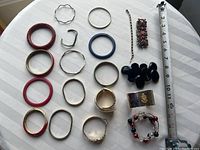 Top-down photo showing all bracelets spread out on a white surface with measuring tape for scale. Includes a variety of metal and colored bangles and a beaded wire bracelet.