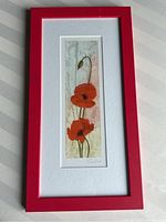 Framed narrow etching print showing two red poppies with stems and bud in front of textured background, framed in red with white mat and artist signature visible.