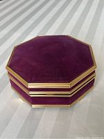 Octagonal purple velvet music jewelry box with gold-metal edge shown closed on white striped fabric.