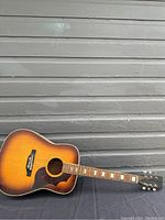 Full view of the acoustic guitar leaning on a wall with sunburst finish, worn and missing strings.