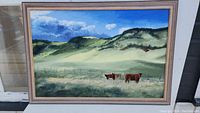 Framed oil painting depicting two cows grazing in a pasture with distant hills and sky.