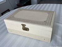 Closed cream-colored rectangular wooden jewelry box with intricate printed border and brass clasp on striped fabric surface, showing slight wear on edges.