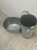 Photo showing two galvanized garden tins on a light wood floor, one tall and cylindrical with embossed design, one oval-shaped