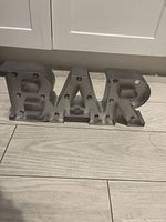 Metal BAR sign with several round bulb sockets on each letter, placed on a light wooden floor in front of white cabinets.