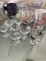 Photo showing various glass stemmed glasses including clear coupe glasses, a purple tinted glass, and decorative wine glass with colorful print. Also included are several tall drinking glasses with red and black print.