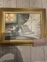 Photo of the framed religious still-life artwork showing a cross, Bible, chalice and grapes in a gold frame on a wood floor