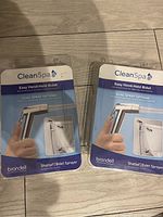 Two unopened packages of Brondell CleanSpa Easy Hand-Held Bidet Sprayers shown side by side on a wooden floor.