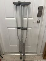 Pair of aluminum underarm crutches with black hand grips and underarm pads, standing against a door.