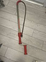 Photo of a manual toilet snake tool with red handles lying on wood floor, showing rust on metal parts.
