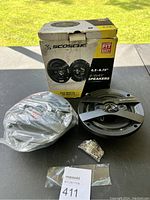 Two Scosche 4-way car speakers with one speaker in plastic wrapping and mounting hardware placed on a flat surface with the original box behind.
