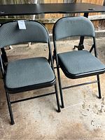 Two gray padded folding chairs with black metal frames standing on concrete floor.