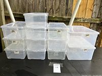 Front view of 14 clear plastic storage bins with lids stacked in three columns of varying sizes on a black surface outdoors.