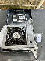 Top view of new Coleman butane stove inside black carrying case, partially wrapped in plastic, showing burner and control knob.