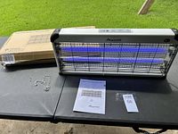 Aspectek bug zapper unit with hanging chain, instruction manual, and original box on display table photographed outdoors with grass background.
