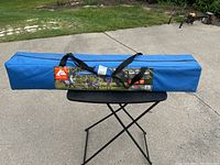 Photo showing the packed blue canopy in its carrying bag with black straps, placed on a folding black table on a driveway, with grass in the background.
