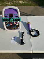 Front and side view of karaoke machine, microphone, and power adapter arranged on white table outdoors.