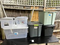 Photo showing clear plastic bins stacked on a black table outside against a wooden fence.