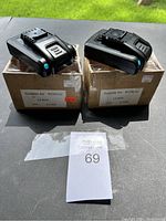 Two black WOX Li-Ion batteries sitting on top of their original brown boxes on outdoor surface with grass background.