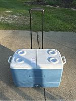 Top view of blue Rubbermaid cooler with white lid and four cup holders, retractable handle extended, sitting on a concrete surface outdoors.