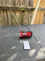 Front and side view of the Midland emergency radio showing the digital display, control buttons, LED flashlight, and extendable antenna on a black outdoor surface.