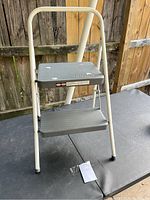 Front angle view of two-step step ladder showing white metal frame and two grey plastic steps on a black surface with wooden fence background.