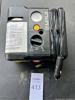 Photo showing black air compressor unit with power cord, pressure gauge, and control dial. Attached to the unit is a power plug designed for a car's 12V outlet. The unit has a label indicating flat tire temporary repair use.