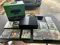 Xbox One console with controller, power adapter, and 10 game cases arranged on table.