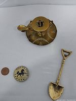 Top view of brass candlestick holder, brass shovel, Combat Dragon II coin, and Elizabeth II 1985 coin with a penny for size comparison.