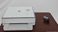White HP Envy 6055 printer shown from angle with extra ink cartridge next to it on table.