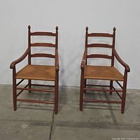 Pair of wooden ladderback chairs with rush seats and armrests, shown from front on concrete floor.