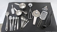 Photo showing full set of metal kitchen utensils and measuring tools arranged on black mat