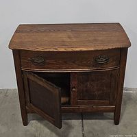 Front view of oak washstand showing curved top, drawer with metal handles, and open left cabinet door revealing interior storage.