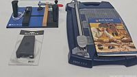 Rockler rail coping sled with two black handles, clamp arm, safety strap, and unopened accessory package. Adjacent to Rockler Edge Pro paper cutter with ruler grid and sliding blade and Rockler instruction manual.